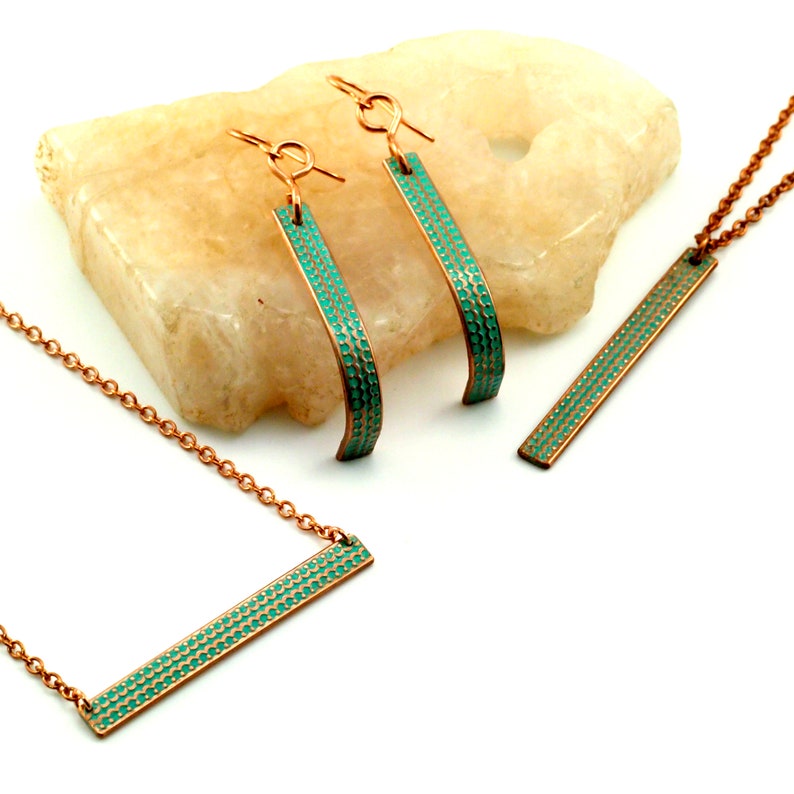 May include: A set of copper jewellery with a turquoise enamel finish. The set includes a necklace, a pair of earrings, and a pendant. The jewellery is displayed on a light brown stone.