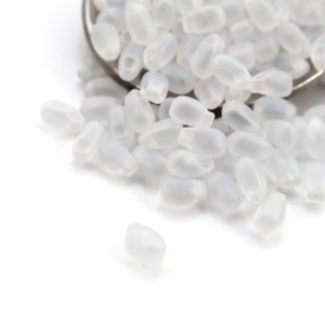 May include: A pile of small, white, opaque glass beads with a slightly iridescent sheen. The beads are oval shaped and have a smooth, frosted finish.