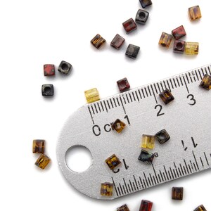 May include: A metal ruler with centimeter markings and a variety of small, square, glass beads in shades of brown, red, and yellow scattered around it.