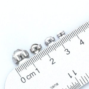 May include: Four silver earring backs of different sizes measured on a ruler. The backs are 0.5 cm, 1 cm, 1.5 cm, and 2 cm in diameter.