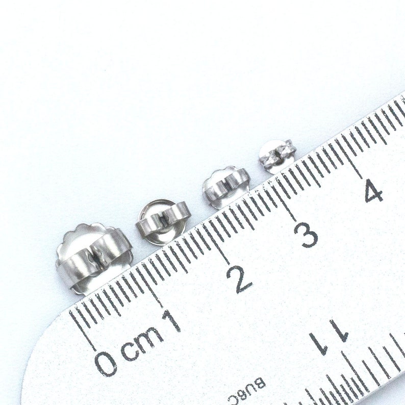May include: Four silver earring backs of different sizes measured on a ruler. The backs are 0.5 cm, 1 cm, 1.5 cm, and 2 cm in diameter.
