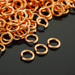 May include: A large collection of small, circular copper-coloured jump rings. Each individual metal ring features a single open cut, allowing for easy connection. The rings have a smooth, polished finish and a consistent round shape throughout.