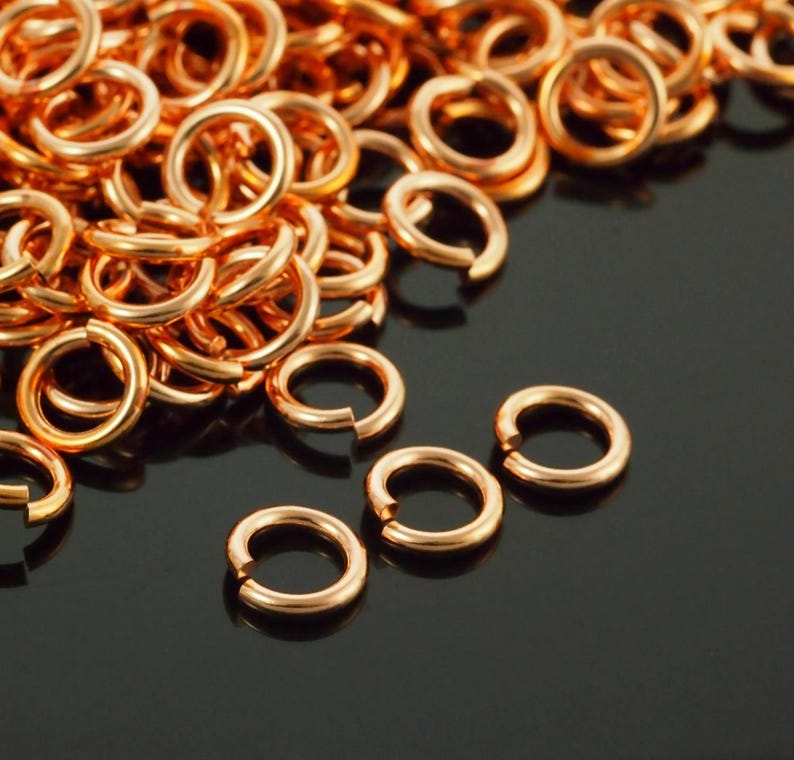 May include: A large collection of small, circular copper-coloured jump rings. Each individual metal ring features a single open cut, allowing for easy connection. The rings have a smooth, polished finish and a consistent round shape throughout.