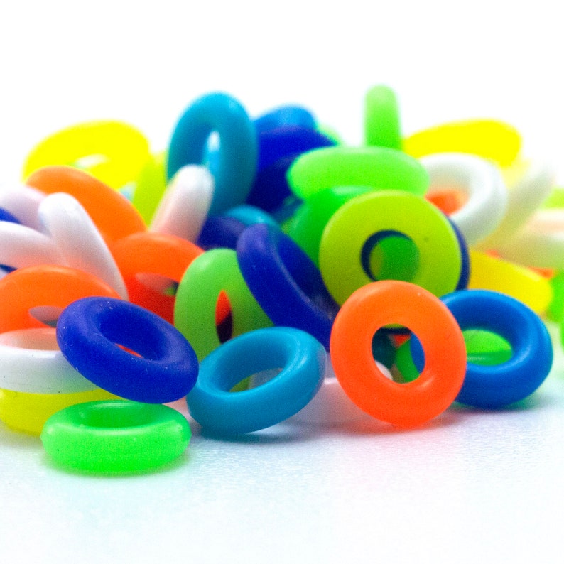 K&ouml;nnte beinhalten: Ein Haufen bunter Silikonringe in verschiedenen leuchtenden Farben, darunter Gr&uuml;n, Blau, Orange, Gelb und Wei&szlig;. Die Ringe sind alle etwa gleich gro&szlig; und in einem zuf&auml;lligen Muster angeordnet.
