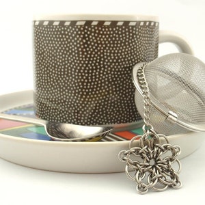Puede incluir: Una taza marr&oacute;n con lunares blancos sobre un plato con un patr&oacute;n geom&eacute;trico colorido. Una cuchara de metal plateado descansa sobre el plato y un infusor de t&eacute; esf&eacute;rico de acero inoxidable con cadena y dije de estrella se sit&uacute;a delante.