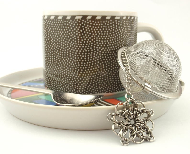 Puede incluir: Una taza marr&oacute;n con lunares blancos sobre un plato con un patr&oacute;n geom&eacute;trico colorido. Una cuchara de metal plateado descansa sobre el plato y un infusor de t&eacute; esf&eacute;rico de acero inoxidable con cadena y dije de estrella se sit&uacute;a delante.