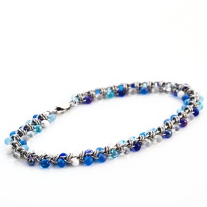 Puede incluir: Una pulsera que presenta una serie de peque&ntilde;as cuentas redondas en varios tonos de azul, blanco y transparente, tejidas juntas con eslabones de alambre de metal plateado. La pieza incluye un cierre de mosquet&oacute;n de metal seguro.