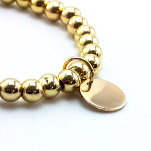 May include: A gold-toned beaded bracelet featuring a series of polished, spherical beads strung together. A small, circular, flat gold-toned charm is attached to the bracelet by a jump ring, hanging from the center of the beaded strand.