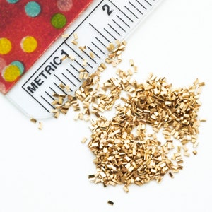 Puede incluir: Primer plano de un mont&oacute;n de confeti met&aacute;lico dorado. El confeti tiene forma de peque&ntilde;os rect&aacute;ngulos. Una regla con la palabra "METRIC" y los n&uacute;meros "1" y "2" es visible en la esquina superior izquierda de la imagen.
