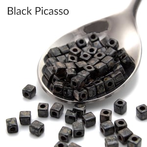 May include: Black Picasso square beads in a silver spoon. The beads are black with white flecks and have a hole in the center. The beads are scattered on a white surface.