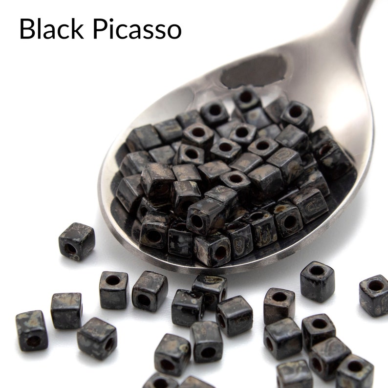 May include: Black Picasso square beads in a silver spoon. The beads are black with white flecks and have a hole in the center. The beads are scattered on a white surface.