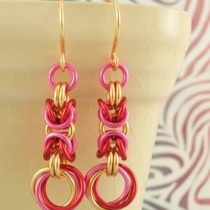 May include: A pair of dangle earrings featuring a chainmaille design with interlocking gold, hot pink, and deep red metal rings. The earrings hang from gold-toned ear wires and display a repeating pattern of circular links in a vertical drop.