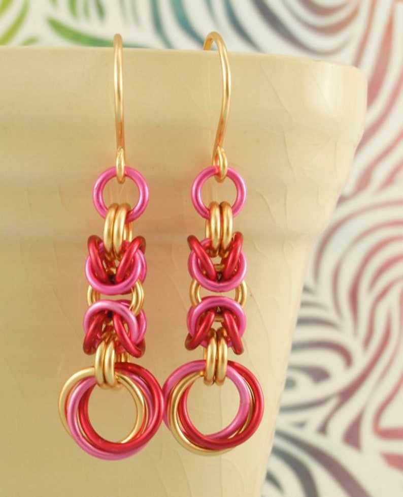 May include: A pair of dangle earrings featuring a chainmaille design with interlocking gold, hot pink, and deep red metal rings. The earrings hang from gold-toned ear wires and display a repeating pattern of circular links in a vertical drop.