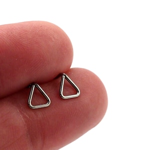May include: Two small, silver-colored triangle-shaped earrings. The earrings are held in a person's fingers against a white background. The earrings are simple and minimalist in design.