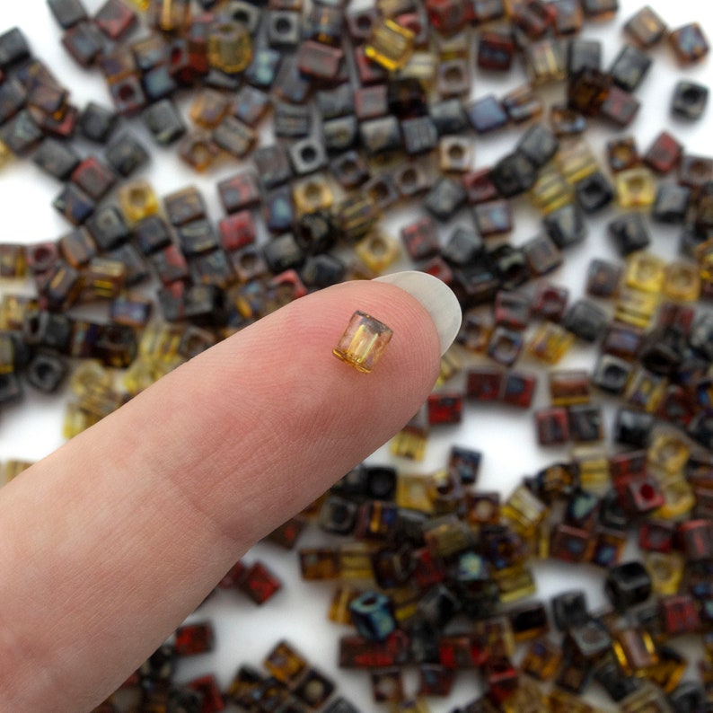 May include: A close-up of a finger holding a small, clear, square-shaped glass bead with a gold tint. The bead is surrounded by a large quantity of similar beads in various colors, including brown, black, and gold.