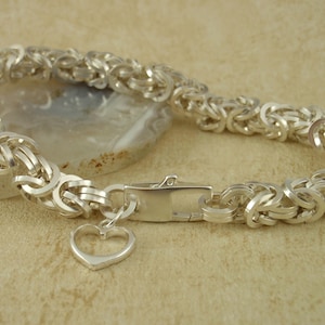 Pode incluir: Uma pulseira prateada com um berloque de coração. A pulseira apresenta um design de corrente trançada e um fecho retangular. O berloque de coração tem um design simples e aberto. A pulseira é exibida em uma superfície marrom clara.