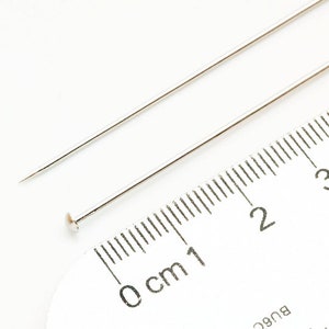 May include: Two silver sewing pins, one with a rounded head, are placed on a white ruler marked in centimeters. The ruler shows measurements from 0 to 3 centimeters.