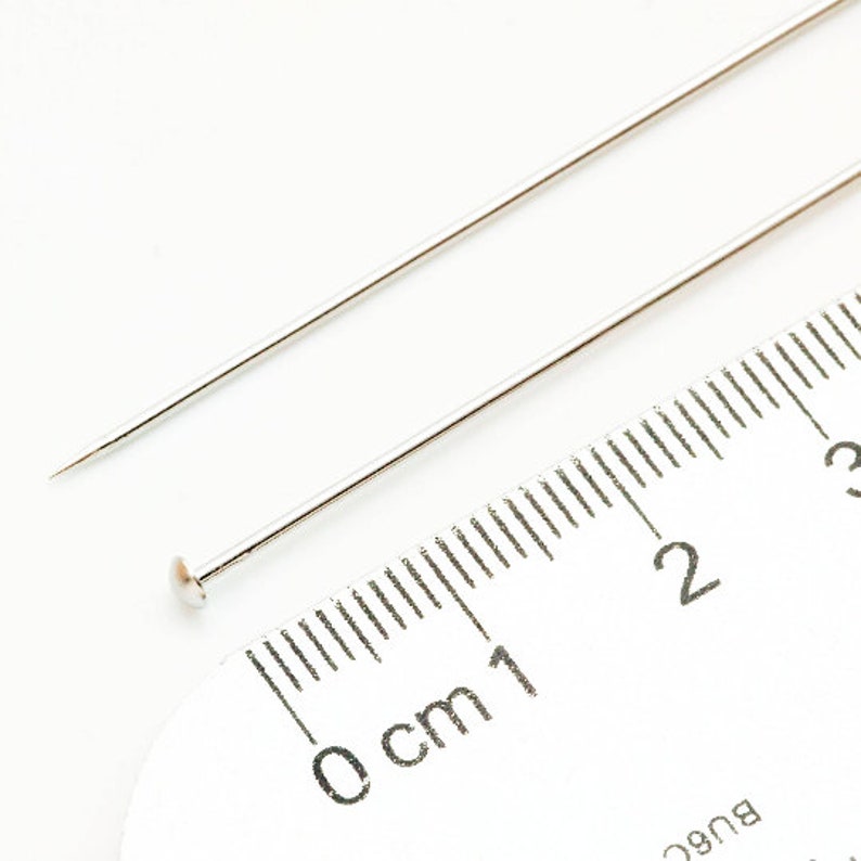 May include: Two silver sewing pins, one with a rounded head, are placed on a white ruler marked in centimeters. The ruler shows measurements from 0 to 3 centimeters.
