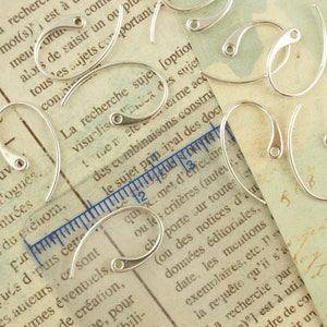 May include: A close-up of a group of silver earring hooks. The hooks are arranged on a piece of paper with text printed on it. The hooks are all the same size and shape, and they have a smooth, polished finish.