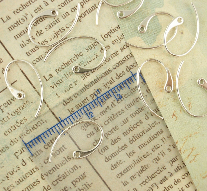 May include: A close-up of a group of silver earring hooks. The hooks are arranged on a piece of paper with text printed on it. The hooks are all the same size and shape, and they have a smooth, polished finish.