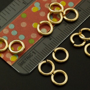 May include: A collection of small, gold-toned metal jump rings scattered near a ruler. The ruler shows both imperial and metric markings, with the rings measuring approximately 0.25 inches in diameter, providing a clear scale for these jewelry components.