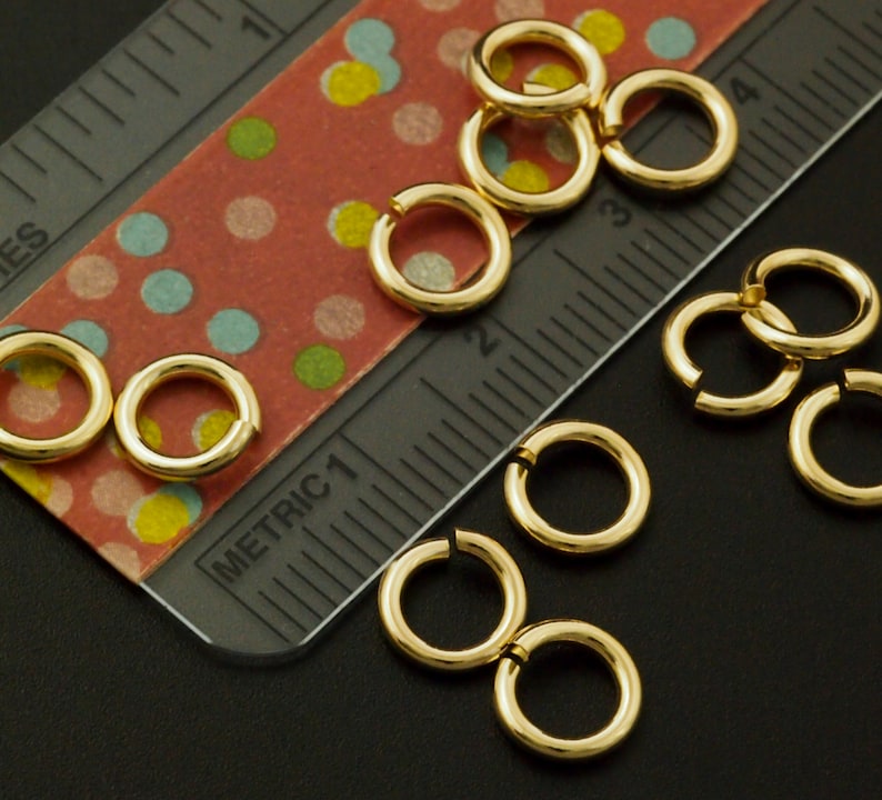 May include: A collection of small, gold-toned metal jump rings scattered near a ruler. The ruler shows both imperial and metric markings, with the rings measuring approximately 0.25 inches in diameter, providing a clear scale for these jewelry components.