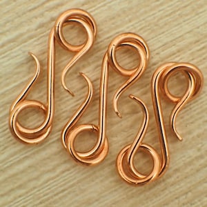 May include: Three copper-coloured metal S-shaped jewellery clasps. Each clasp features a continuous wire design with two circular loops at opposite ends, connected by a curved central bar. The metal has a smooth, polished finish with a warm metallic tone.