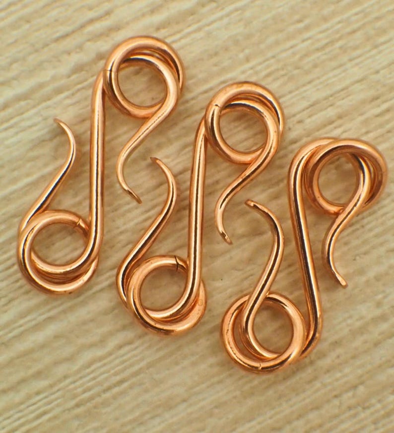 May include: Three copper-coloured metal S-shaped jewellery clasps. Each clasp features a continuous wire design with two circular loops at opposite ends, connected by a curved central bar. The metal has a smooth, polished finish with a warm metallic tone.