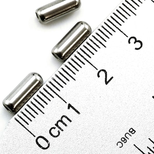 May include: Three silver metal tubes, each measuring approximately 1 cm in length, are placed on a ruler with markings in centimeters. The ruler shows measurements from 0 cm to 3 cm.