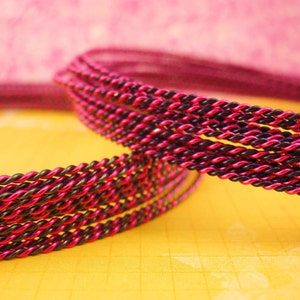 K&ouml;nnte beinhalten: Eine gedrehte Kordel mit zweifarbigem Design aus abwechselnden Str&auml;ngen in leuchtendem Pink und tiefem Schwarz. Die Kordel ist in mehreren Schlaufen gewickelt und zeigt die gleichm&auml;&szlig;ige, fest gewundene Textur der metallisch wirkenden Fasern.