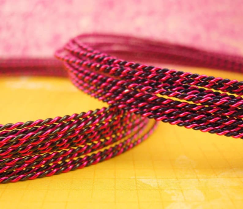 K&ouml;nnte beinhalten: Eine gedrehte Kordel mit zweifarbigem Design aus abwechselnden Str&auml;ngen in leuchtendem Pink und tiefem Schwarz. Die Kordel ist in mehreren Schlaufen gewickelt und zeigt die gleichm&auml;&szlig;ige, fest gewundene Textur der metallisch wirkenden Fasern.