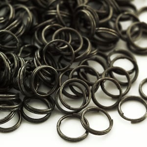 May include: A collection of dark gunmetal gray split rings, each featuring a double-loop coil design. These small, circular metal findings are constructed from thin wire and are arranged in a loose, overlapping pile with a metallic finish.