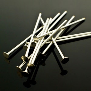 May include: A pile of silver-toned head pins with flat round heads. The pins are arranged in a random pattern on a black surface.