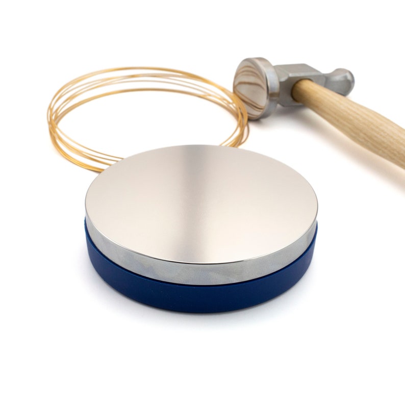 May include: A jewelry-making setup featuring a silver disc with a blue base, a small hammer with a wooden handle, and a coil of gold wire. The tools are arranged on a white background, suggesting a craft or hobby.