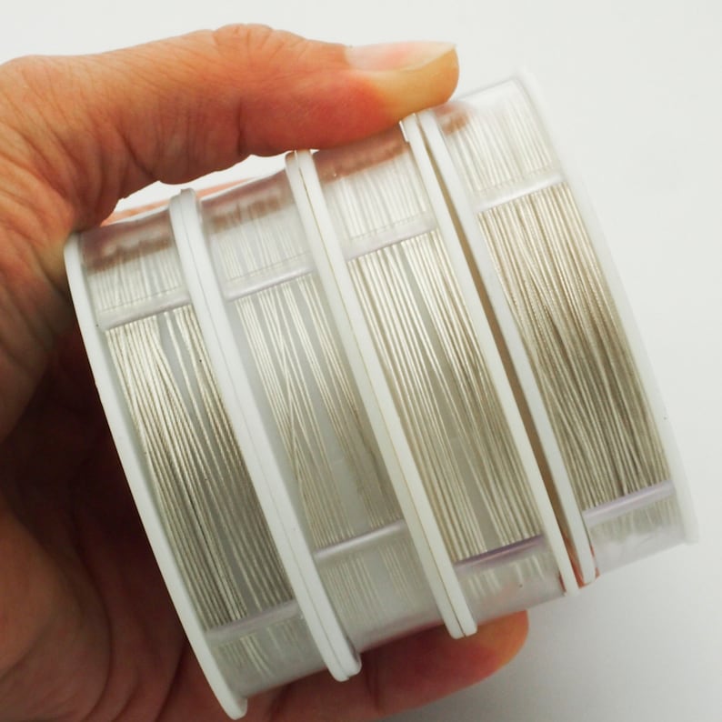 Puede incluir: Cuatro carretes de pl&aacute;stico transparente apilados, cada uno envuelto con una longitud continua de alambre met&aacute;lico fino de color plateado. Los carretes cuentan con tapas circulares blancas y est&aacute;n dispuestos horizontalmente.