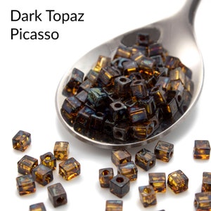 May include: A close-up of a spoon filled with dark topaz Picasso square beads. The beads are scattered on a white surface.