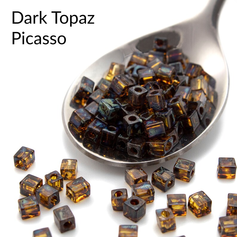 May include: A close-up of a spoon filled with dark topaz Picasso square beads. The beads are scattered on a white surface.