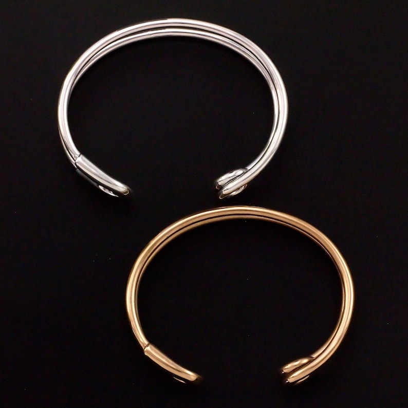 K&ouml;nnte beinhalten: Zwei schlichte Armreifen, einer in Silber und einer in Gold, mit einem Knotendesign am Verschluss.