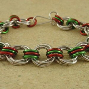 May include: A chainmail bracelet constructed from interlocking silver, red, and green metal rings. The bracelet features a repeating pattern of these colored rings linked together and includes a simple silver metal hook clasp for closure.