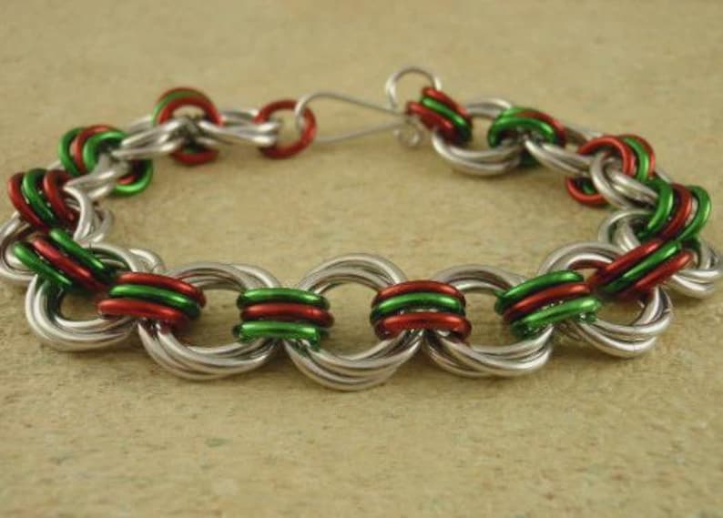 May include: A chainmail bracelet constructed from interlocking silver, red, and green metal rings. The bracelet features a repeating pattern of these colored rings linked together and includes a simple silver metal hook clasp for closure.