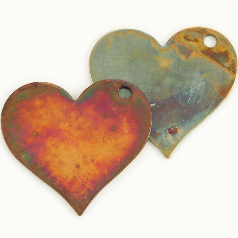May include: Two heart-shaped metal charms with a distressed, oxidized finish. One charm is copper-toned with a reddish hue, while the other is a silvery-green color. Both charms have a small hole at the top for attaching to jewelry or other crafts.