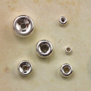 May include: Six silver metal donut-shaped beads with a hole in the centre. The beads are different sizes and are arranged in a random pattern on a beige surface.