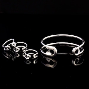 K&ouml;nnte beinhalten: Ein Set silberner Sicherheitsnadelringe und ein silbernes Sicherheitsnadel-Armband. Die Ringe sind in verschiedenen Gr&ouml;&szlig;en und das Armband ist offen und hat einen Sicherheitsnadelverschluss.