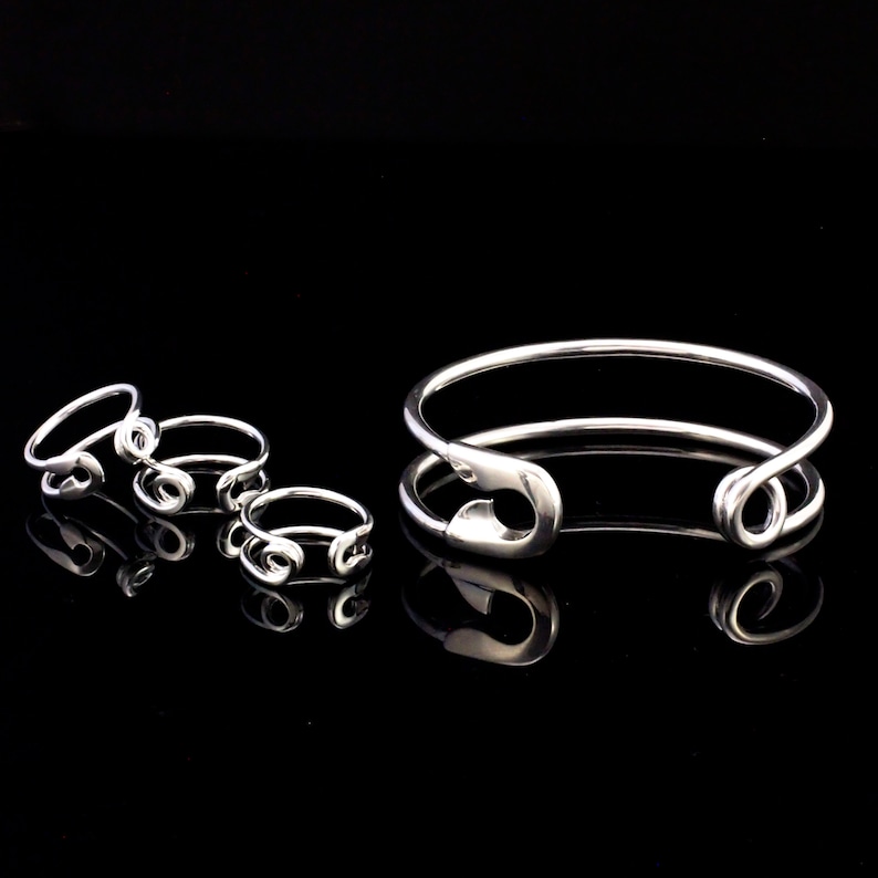 K&ouml;nnte beinhalten: Ein Set silberner Sicherheitsnadelringe und ein silbernes Sicherheitsnadel-Armband. Die Ringe sind in verschiedenen Gr&ouml;&szlig;en und das Armband ist offen und hat einen Sicherheitsnadelverschluss.