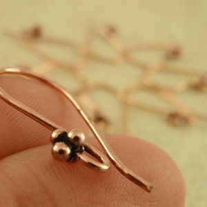 May include: A close-up of a copper earring hook with three small round beads at the end. The hook is made of a thin wire and is designed to be worn in the ear.