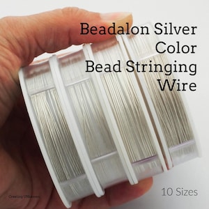 Puede incluir: Carretes de hilo para ensartar cuentas Beadalon color plata. El hilo está enrollado firmemente en carretes de plástico blanco. El texto de la imagen dice "Beadalon Silver Color Bead Stringing Wire" y "10 Sizes".