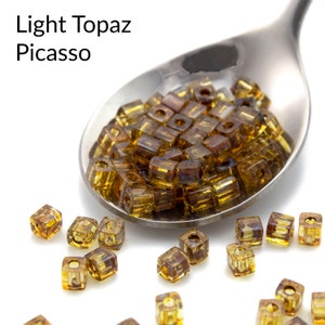 May include: A spoon filled with light topaz Picasso square glass beads. The beads are a light brown color with a slight shimmer. There are also several beads scattered on a white surface.
