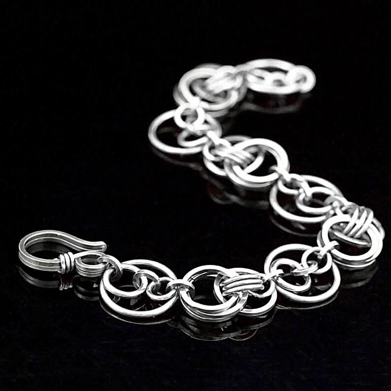 May include: A silver bracelet composed of linked circular elements. The bracelet has a hook clasp. The design is simple and elegant, suitable for everyday wear. The bracelet is displayed against a black background. The bracelet's length is approximately 18 cm.