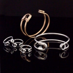 K&ouml;nnte beinhalten: Ein Set aus drei silbernen Sicherheitsnadelringen und zwei silbernen Sicherheitsnadelarmb&auml;ndern, eines davon mit einem goldenen Sicherheitsnadelarmband.