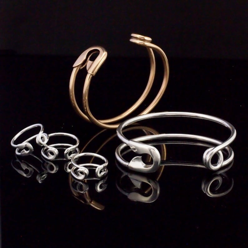 K&ouml;nnte beinhalten: Ein Set aus drei silbernen Sicherheitsnadelringen und zwei silbernen Sicherheitsnadelarmb&auml;ndern, eines davon mit einem goldenen Sicherheitsnadelarmband.